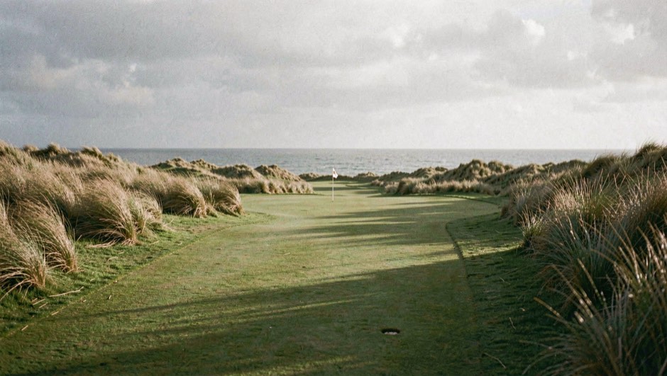 Golfbaan in de duinen bij laag ochtendlicht, met een gemaaide fairway die tussen helmgras naar een verre vlag loopt